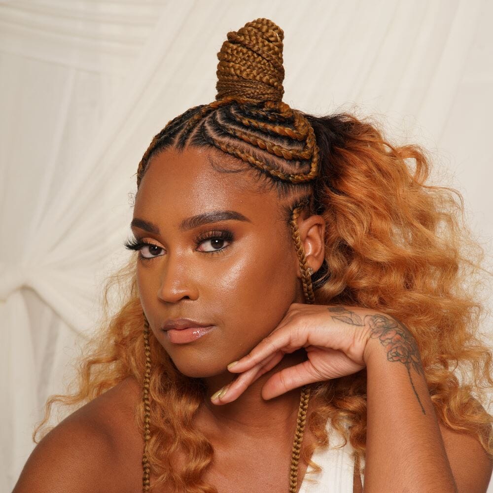 A woman with a half-updo with braids wrapped into a topknot. She looks at the camera with a hand under her face.