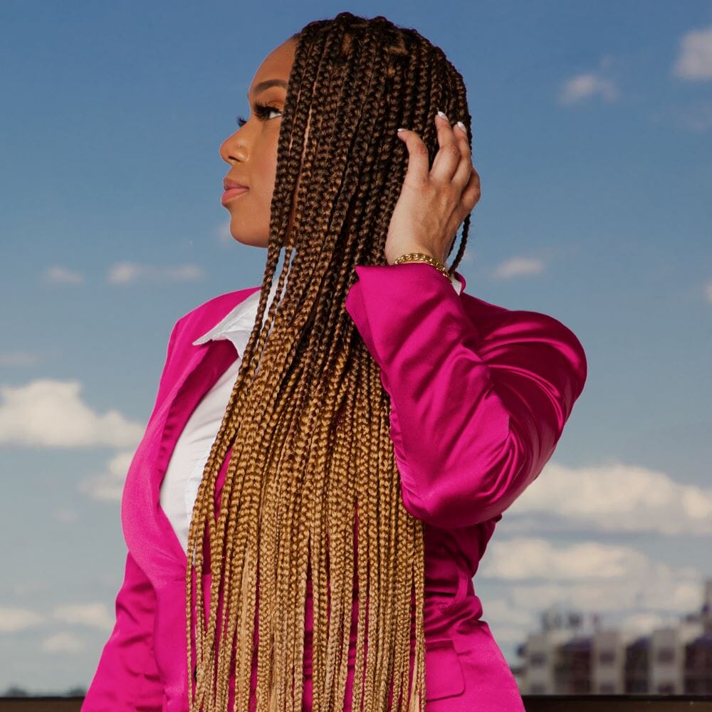 A woman in a pink blazer stands looking away from the camera. She has long brown-gold ombré braids.