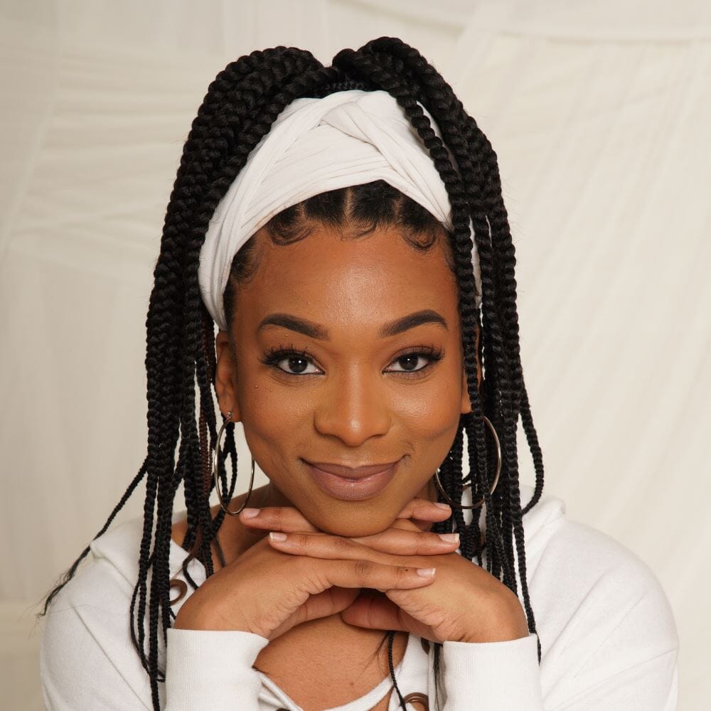 Close-up of a woman with her hair up in long black box braids wrapped in a white headband. She is resting her chin on her hands.