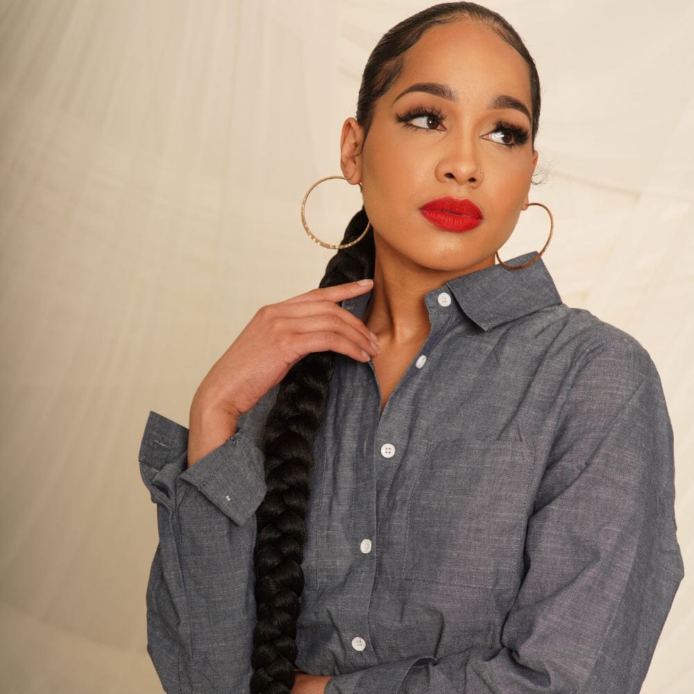 A woman in a button-down, red lips and gold hoops stands resting her hand on her long braid of black hair.