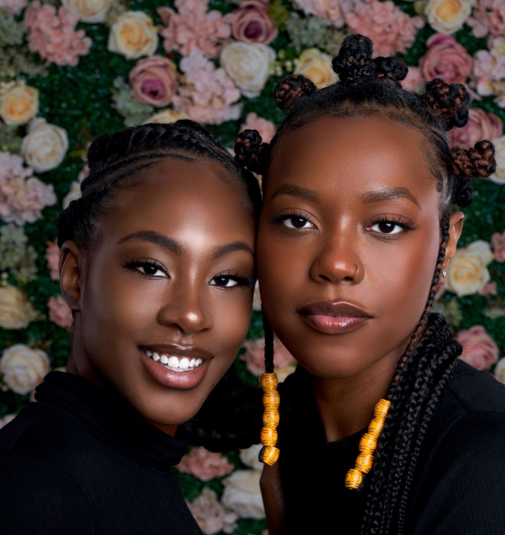 Close-up portrait of two black women with braided hairstyles, standing in front of a floral background.