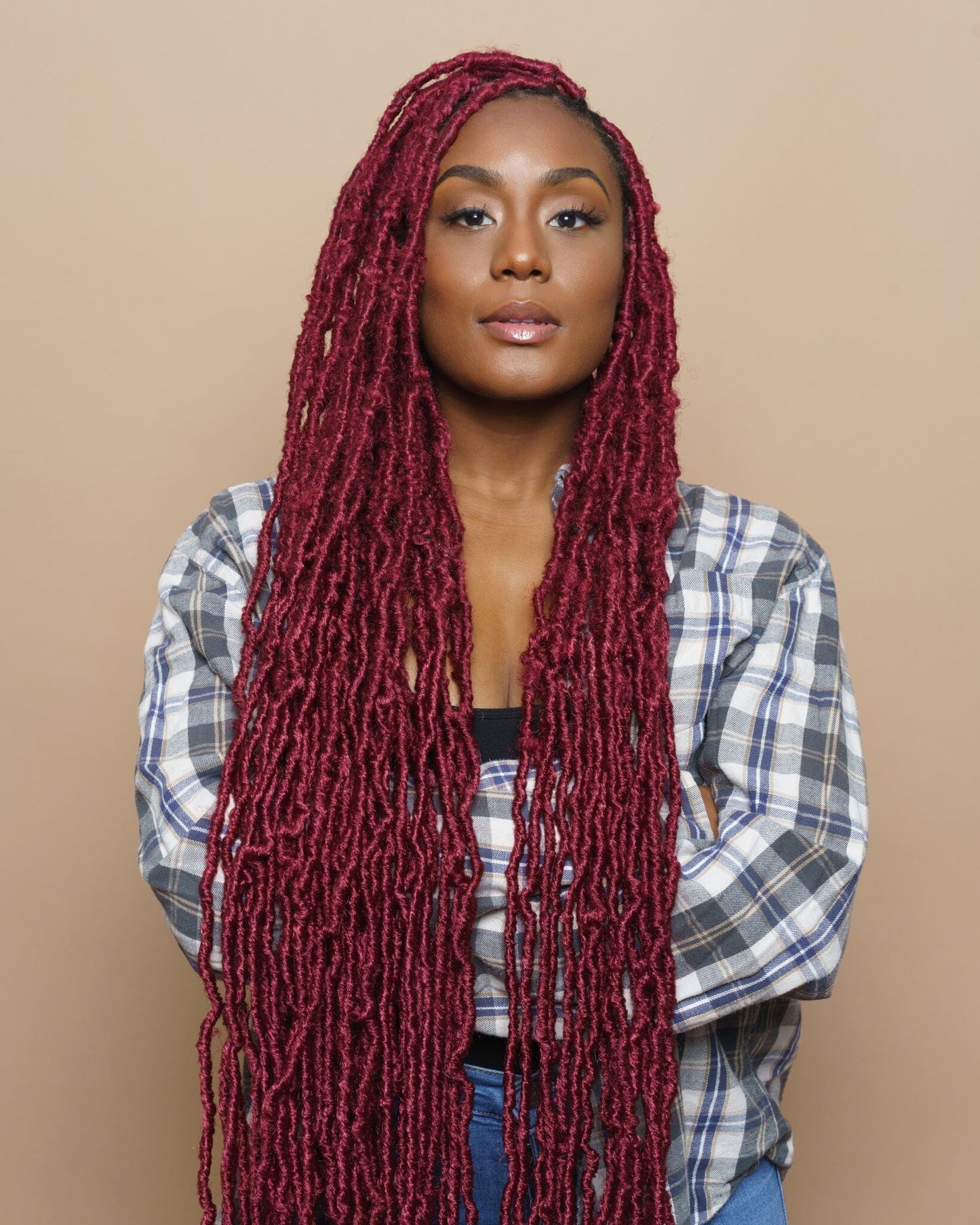 A black woman with long, dark red locs stands with her arms folded, looking at the camera. She wears a plaid shirt and jeans.